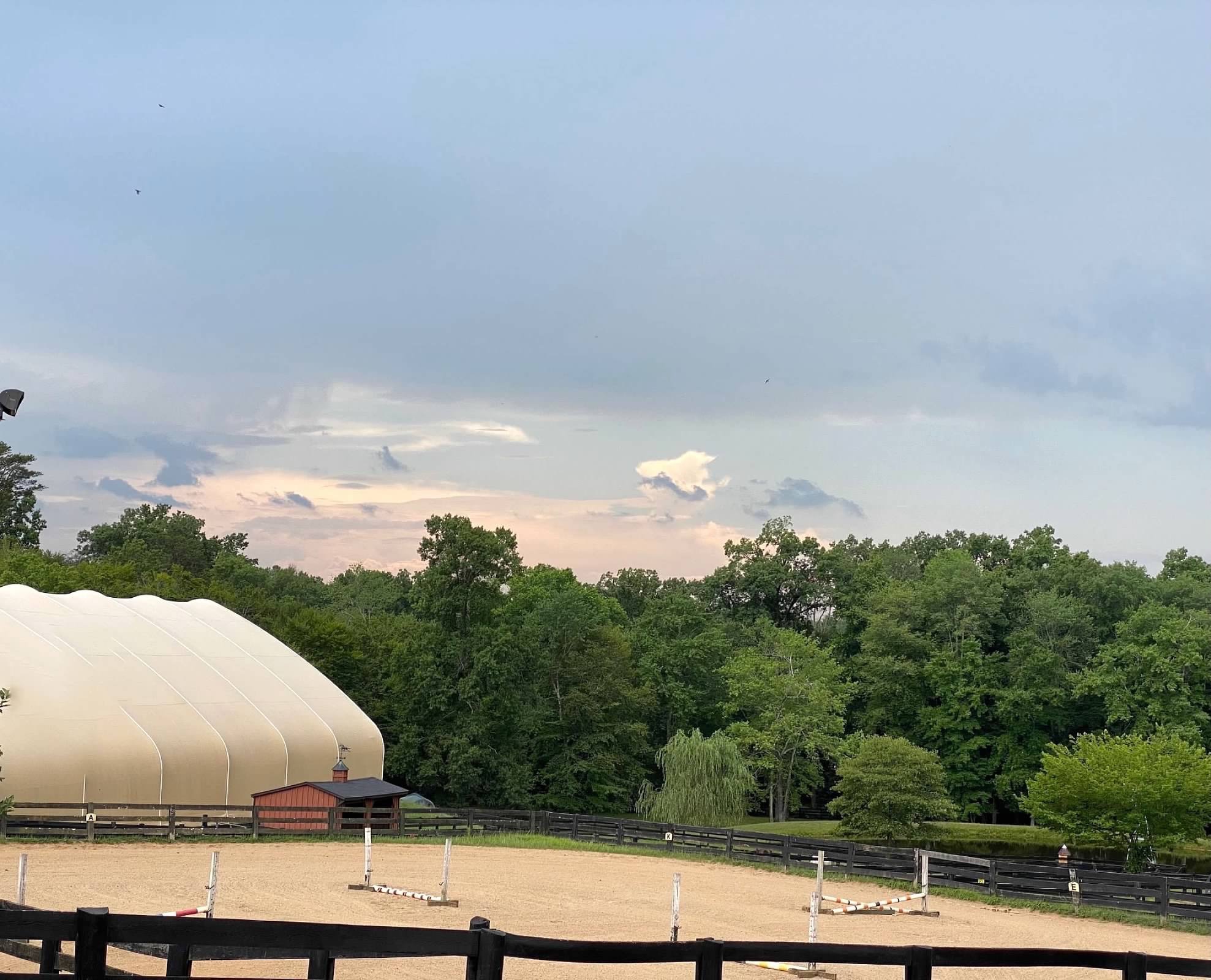 The covered arena beside the outdoor sand ring with jumps set on a clear summer evening