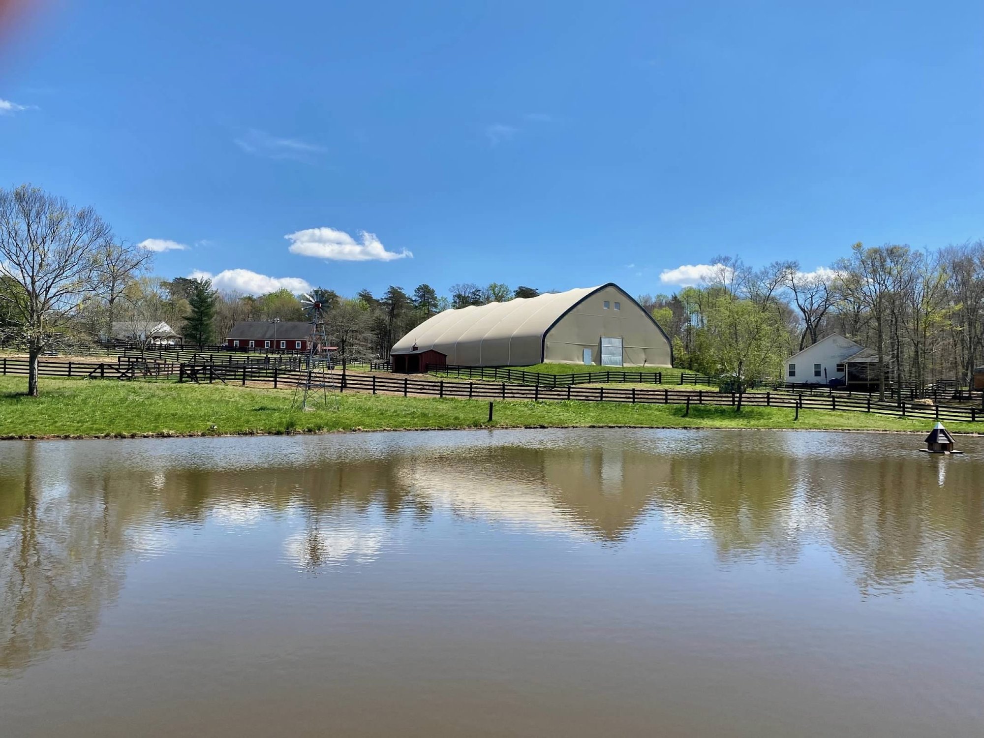 The Brentsville Stables property seen across the pond — covered arena, red barn, windmill, and farmhouse under a blue sky
